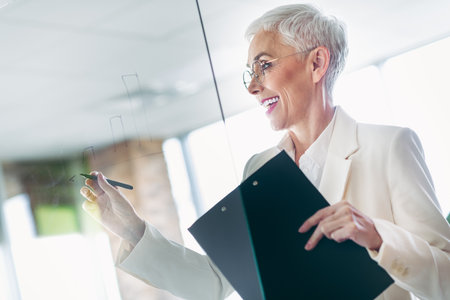 Senior businesswoman presenting data insights on a glass board in an office setting, demonstrating professionalism and confidenceの写真素材