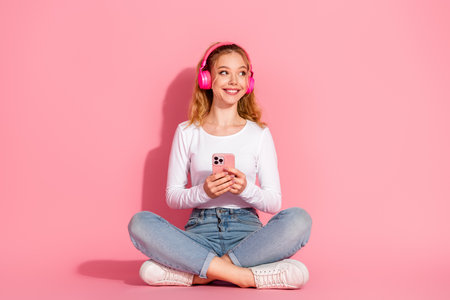 Charming young girl listening to music with headphones seated cross-legged on a pink background and holding a smartphoneの写真素材