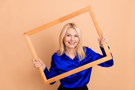 Smiling mature woman holding a wooden frame, expressing creativity and positivity against a beige...