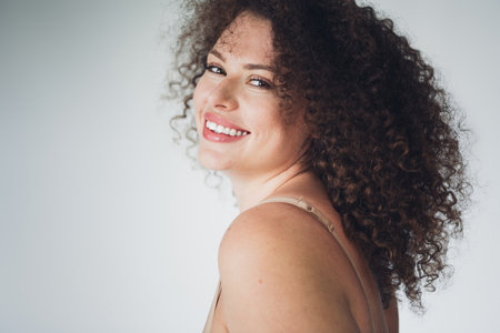 Charming woman with curly hair smiling in natural beauty on a soft white background, celebrating body positivity and self acceptance.の写真素材