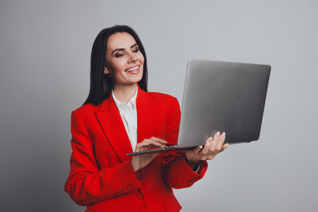 Confident young businesswoman in a stylish red suit using a laptop against a grey background, showcasing professionalism and eleganceの写真素材
