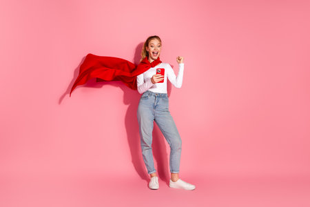 Young cheerful woman in casual style gesturing with exciting energy while holding a smartphone against a lively pink backdropの写真素材