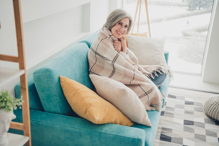 Charming mature woman relaxing on a cozy sofa with warm blanket at home by a bright window on a snowy dayの写真素材