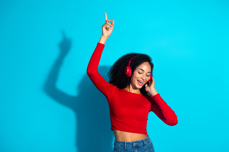 Young stylish woman enjoying music with headphones on a vibrant blue background, wearing a red top,...