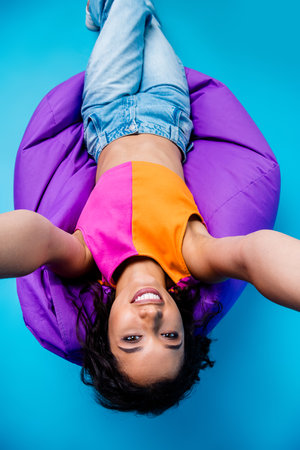 Young woman with vibrant outfit relaxing on a purple bean bag against a bright blue background, smiling confidentlyの写真素材