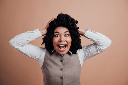 Excited businesswoman with curly hair holding head with both hands against beige background expressing joy and surpriseの写真素材