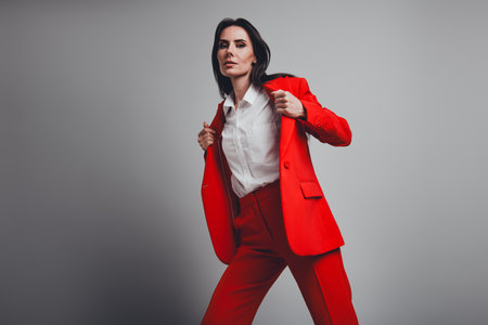 Successful brunette woman in elegant red suit posing against grey background expressing authority and stylish charmの写真素材