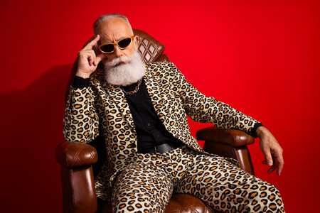 Charismatic elderly gentleman with a stylish leopard print suit posing confidently in a chair against a red background.の写真素材