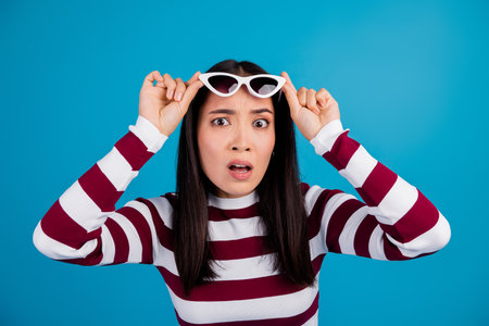 Portrait of a surprised young woman adjusting sunglasses against a blue background wearing a striped shirtの写真素材
