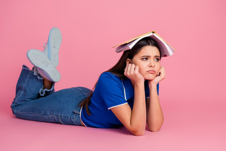 Young female student lying on pink background with book on head, showing expressions of frustrationの写真素材