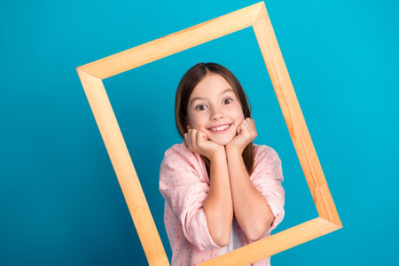 Cheerful young girl posing with a wooden frame on a vibrant blue background, wearing a casual pink shirtの写真素材
