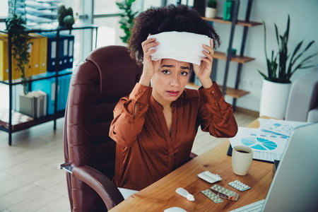 Female entrepreneur distressed in workplace managing health while balancing professional duties inside a chic modern office setup indoorsの写真素材