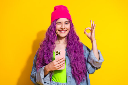 Stylish young woman with purple hair showing an OK hand gesture while holding a smartphoneの写真素材