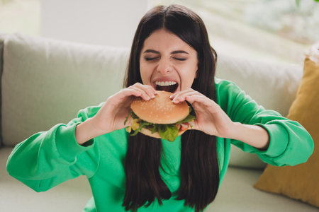 Young brunette woman enjoying a tasty burger at home in casual green pullover, sitting on a sofa in...