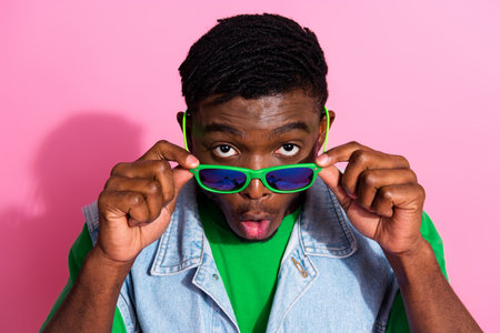 Trendy young man in denim vest making a playful gesture with stylish sunglasses against a vibrant...
