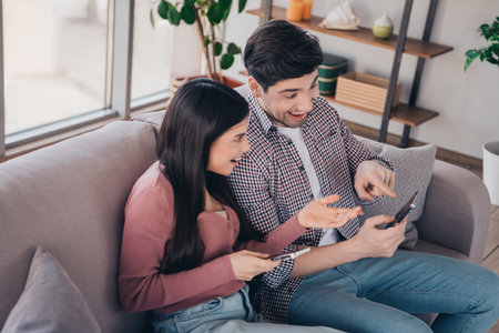 Cheerful young couple enjoying time together at home sitting on a couch sharing laughs while browsing smartphonesの写真素材