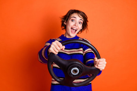 Photo portrait of young girl wearing striped blue sweater holding steering wheel driving first vehicle isolated on orange color backgroundの写真素材