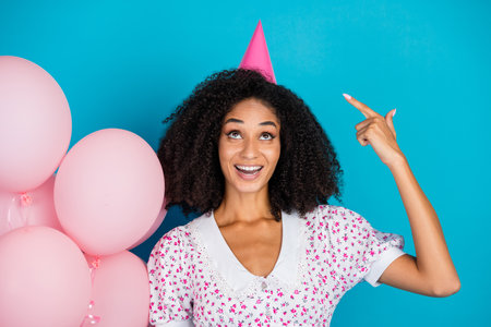 Smiling young woman wearing a floral dress celebrates with pink balloons against a bright blue...
