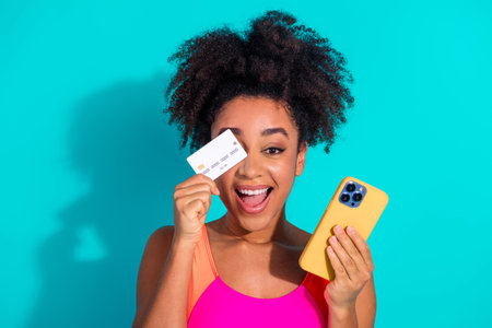 Smiling young woman holding a credit card and smartphone against a vibrant turquoise background showcasing excitementの写真素材