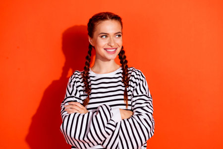 Young woman with red hair wearing a striped pullover stands confidently against a vibrant orange background, exuding happiness and charmの写真素材