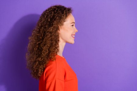 Side profile view of a cheerful young woman with curly hair wearing an orange top against a vivid purple backgroundの写真素材