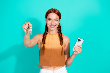 Smiling young woman with red braided hair holding house keys and a smartphone on a turquoise background, symbolizing new ownershipの写真素材