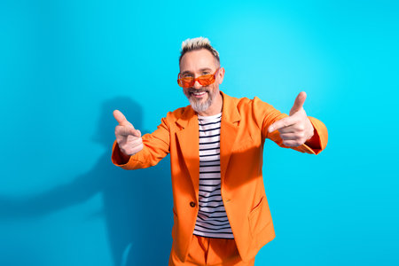 Charismatic middle aged man in orange suit gesturing confidently against vibrant blue background, exuding elegance and styleの写真素材