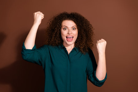 Photo of cheerful lucky lady dressed green shirt rising fists empty space isolated brown color backgroundの写真素材