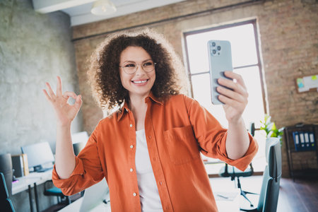 Confident Young Businesswoman Taking a Selfie in a Modern Office Environment While Expressing Positivity and Successの写真素材
