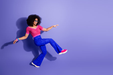 Stylish young woman in trendy colorful outfit posing against purple background, reflecting retro...