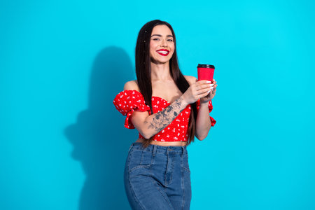 Young woman holding a pink coffee cup in front of a vibrant blue background, wearing a red top and jeans, smiling confidentlyの写真素材