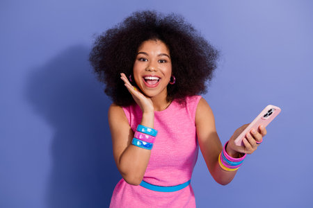 Trendy young woman with curly hair joyfully posing in stylish pink dress against a vibrant purple...