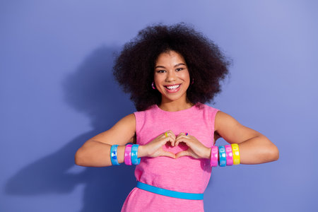 Smiling young woman in retro fashion style making a hand heart gesture against a purple backdrop in...