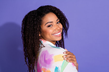 A young woman with curly hair and a vibrant shirt smiles brightly against a purple background,...