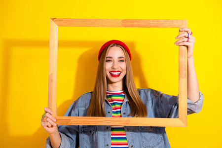 Smiling young woman holding a wooden frame, wearing casual denim and a red beanie, with a bright...