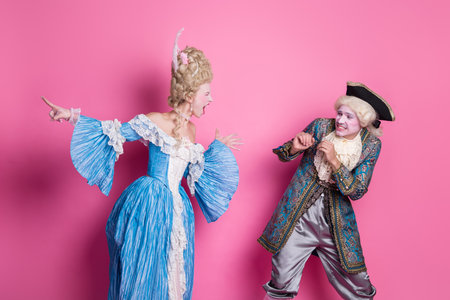 A couple in baroque costumes shares a playful moment against a vibrant pink backdrop, embodying elegance and humorの写真素材