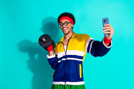 Young man in retro sportswear smiling while holding dumbbell and taking smartphone selfie on a vibrant teal studio backgroundの写真素材