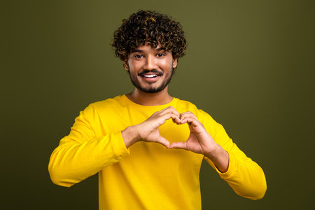 Young man in yellow making heart hand gesture on khaki background for cheerful concepts and lifestyle campaignsの写真素材