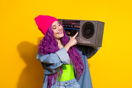 Stylish young woman holding a boombox against a vivid yellow background wearing casual denim, with...