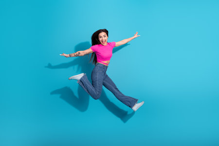 Energetic young woman jumping joyfully wearing a pink top and blue jeans on a vibrant blue...