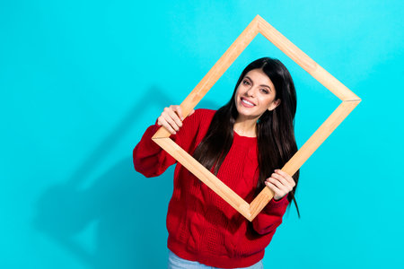 Smiling woman holding a wooden frame and wearing a red sweater, standing against a bright turquoise teal backgroundの写真素材