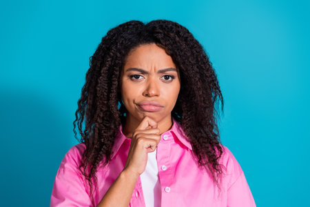 Thoughtful young woman with curly hair in a pink shirt contemplating an idea against a vibrant blue...