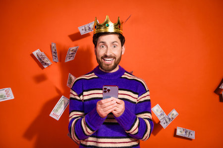 Photo of attractive young man amazed falling money hold device dressed stylish striped purple clothes isolated on orange color backgroundの写真素材
