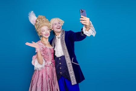 Playful couple dressed in elegant baroque costumes taking a selfie against a vibrant blue backgroundの写真素材