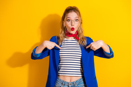Blond Woman in Blue Jacket and Red Scarf Against Bright Yellow Background, Pointing at Herselfの写真素材