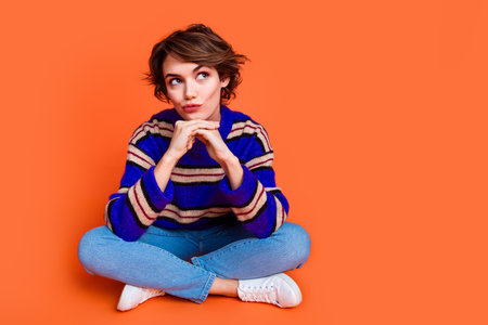 Photo portrait of young brown hair lady in blue striped cute sweater sitting floor deep thinking pensive isolated on orange color backgroundの写真素材