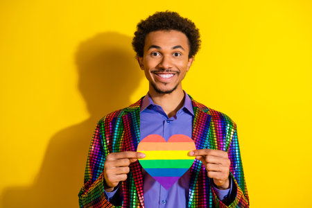 Vibrant young man holding a rainbow heart symbol against a bright yellow background, showcasing diversity, prideの写真素材