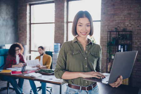 Young confident professional using a laptop in a relaxed modern coworking office with colleagues collaborating in the backgroundの写真素材