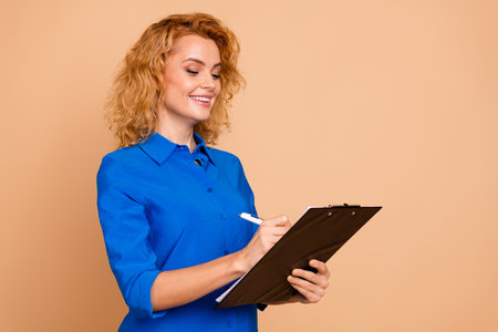 Confident businesswoman in a blue shirt smiling and jotting notes on a clipboard with a professional...