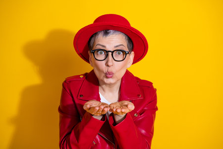 Fashionable elderly woman in red leather jacket and hat gesturing with hands against bright yellow backgroundの写真素材